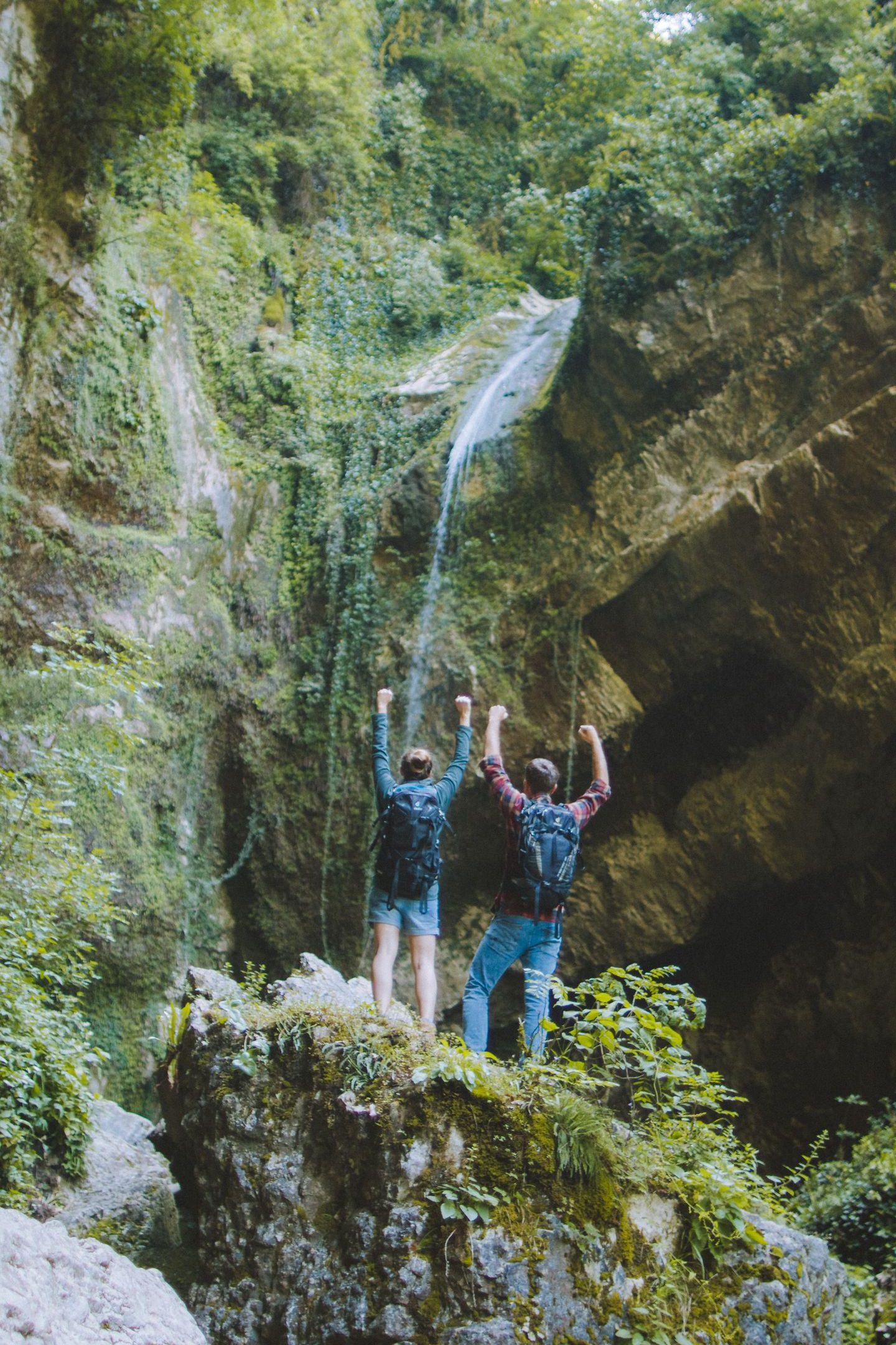 A couple of hikers raise their arms victoriously on a mossy rock near a scenic waterfall in a lush forest.